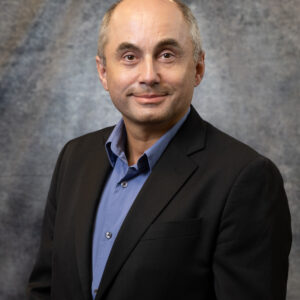 Studio portrait of Burak Ozpineci, Section Head for Vehicle and Mobility Systems at Oak Ridge National Laboratory, wearing a black jacket and blue shirt while facing the camera against a gray textured backdrop at the National Transportation Research Center.