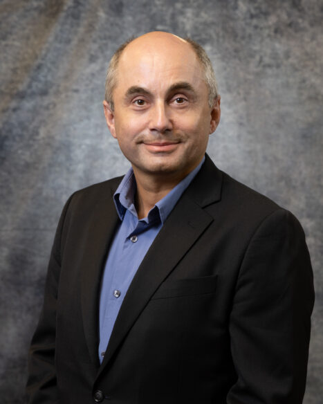 Studio portrait of Burak Ozpineci, Section Head for Vehicle and Mobility Systems at Oak Ridge National Laboratory, wearing a black jacket and blue shirt while facing the camera against a gray textured backdrop at the National Transportation Research Center.