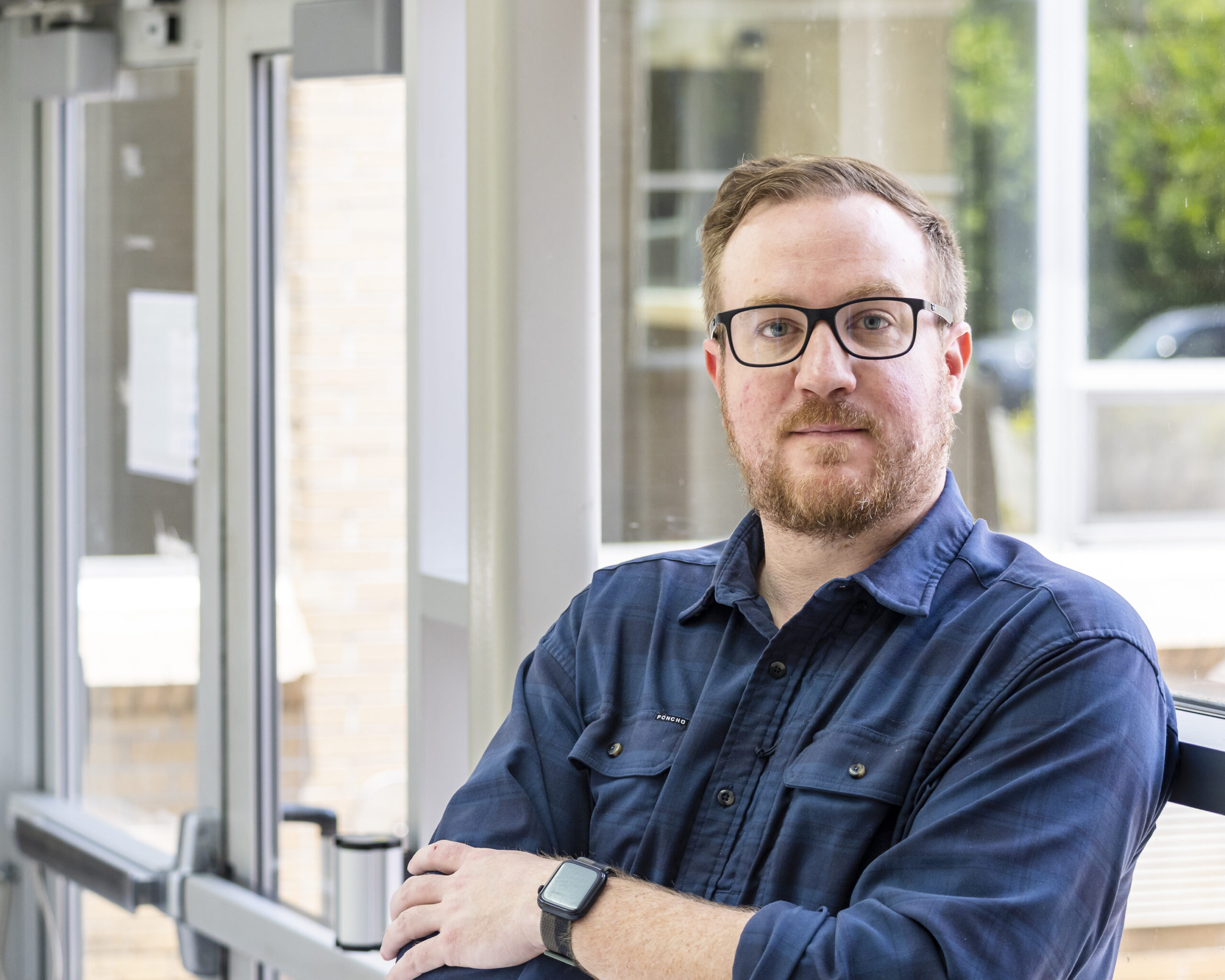 Man is leaning against the window, arms crossed in a dark navy button up. 