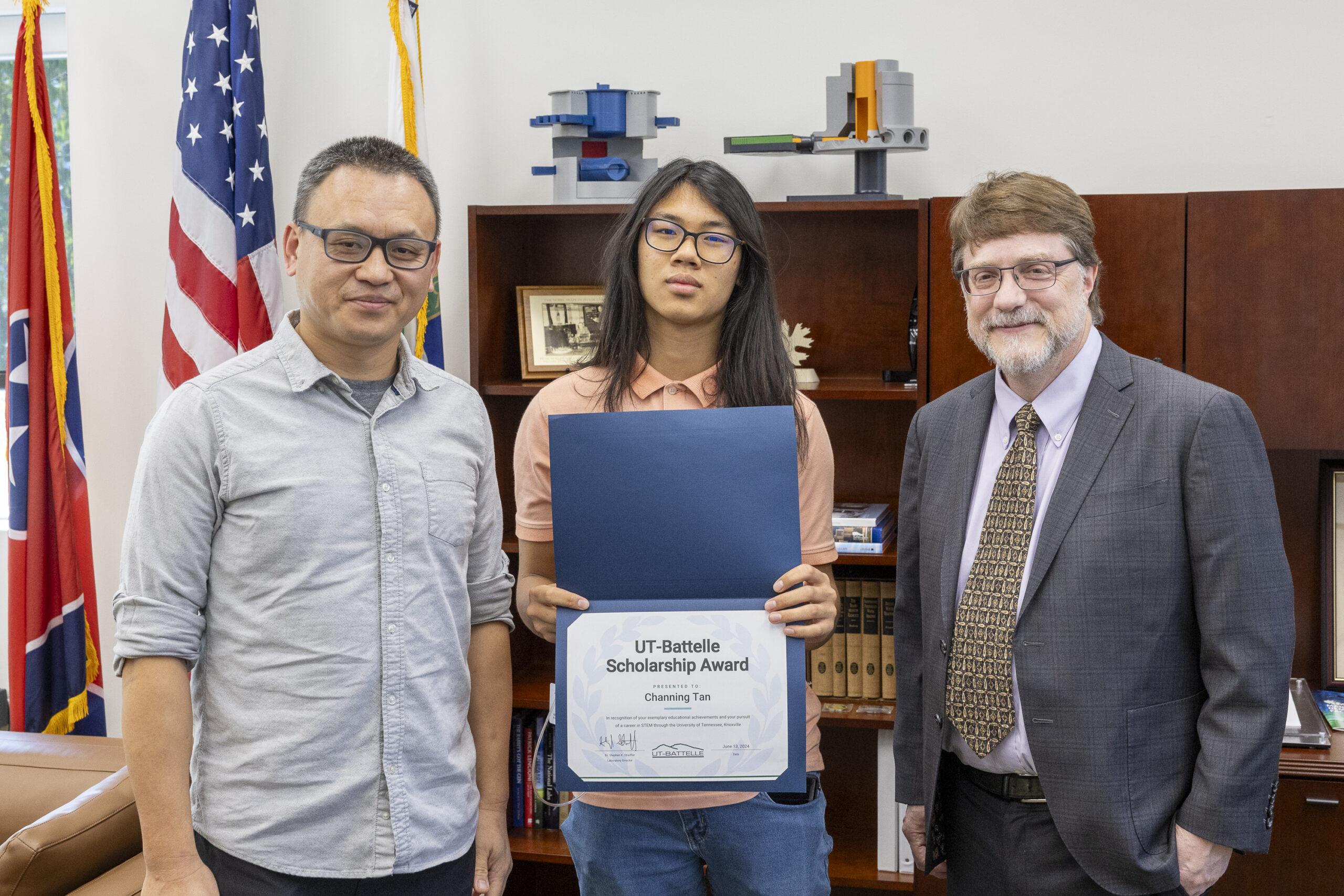 Three people are standing in an office with the middle person holding an award. 