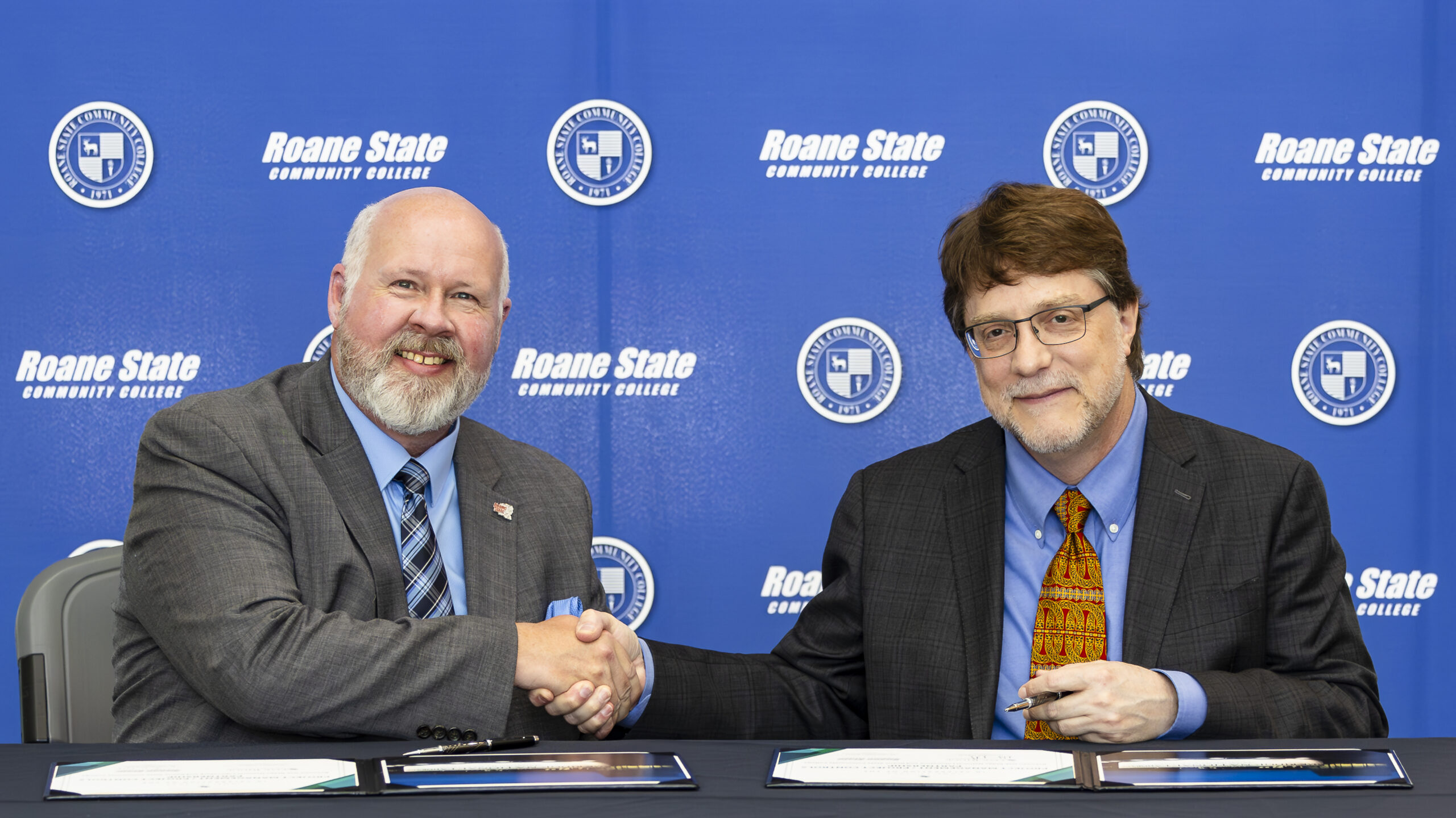 Two men shake hands in front of a blue background. Both are wearing suites and a tie.