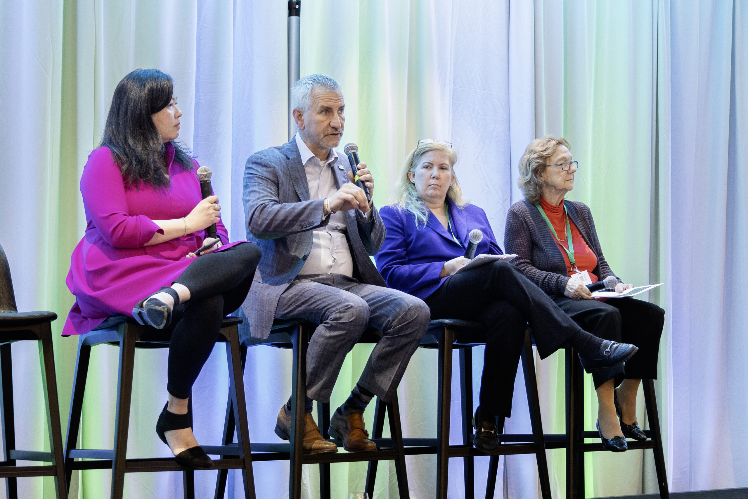 Four panelists sit on tall chairs on a stage with green and blue background
