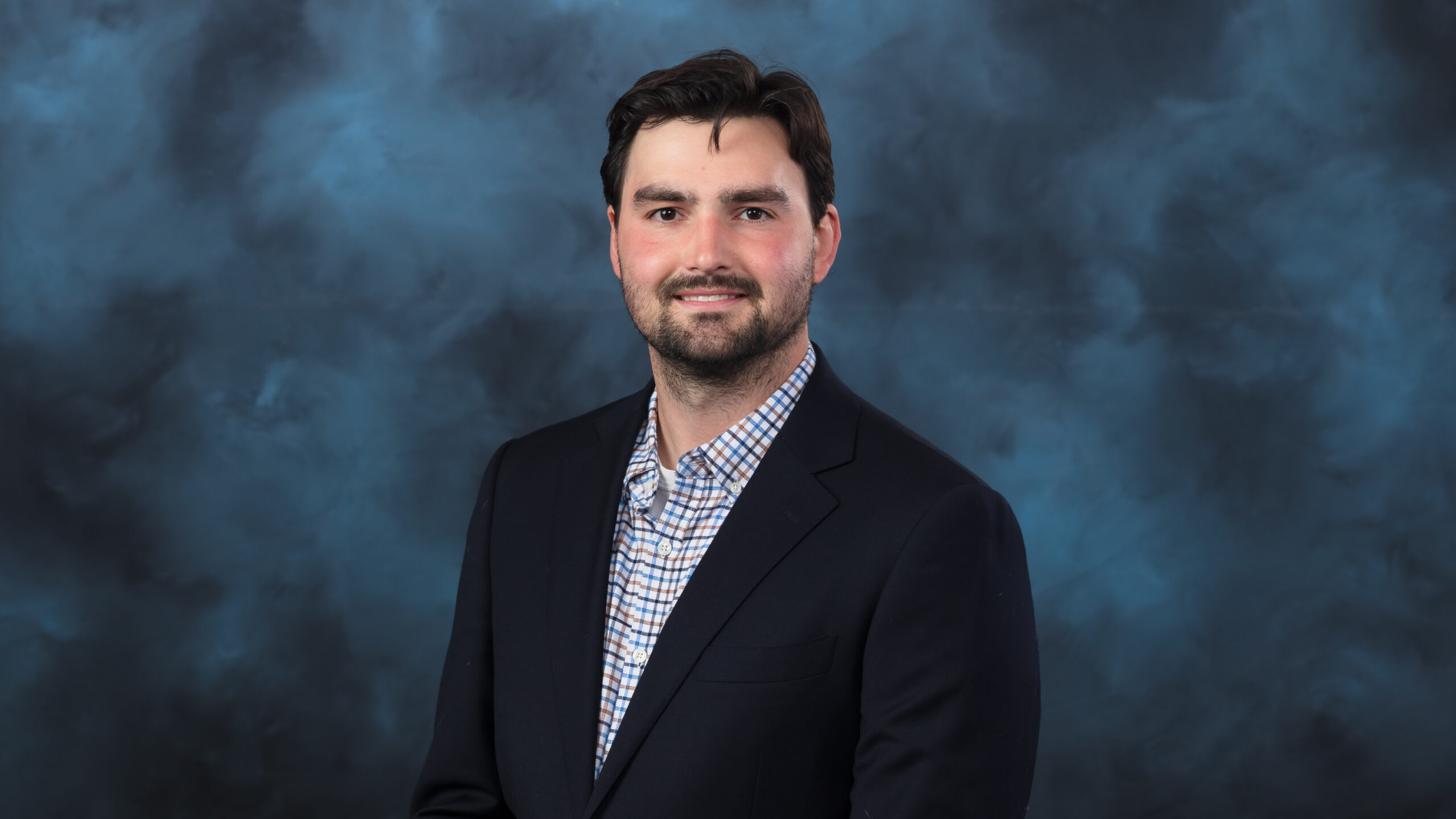 Alec Readel, Innovation Crossroads, portrait. Young man with a beard and wearing a suit jacket sits in front of a blue background for a formal photo.