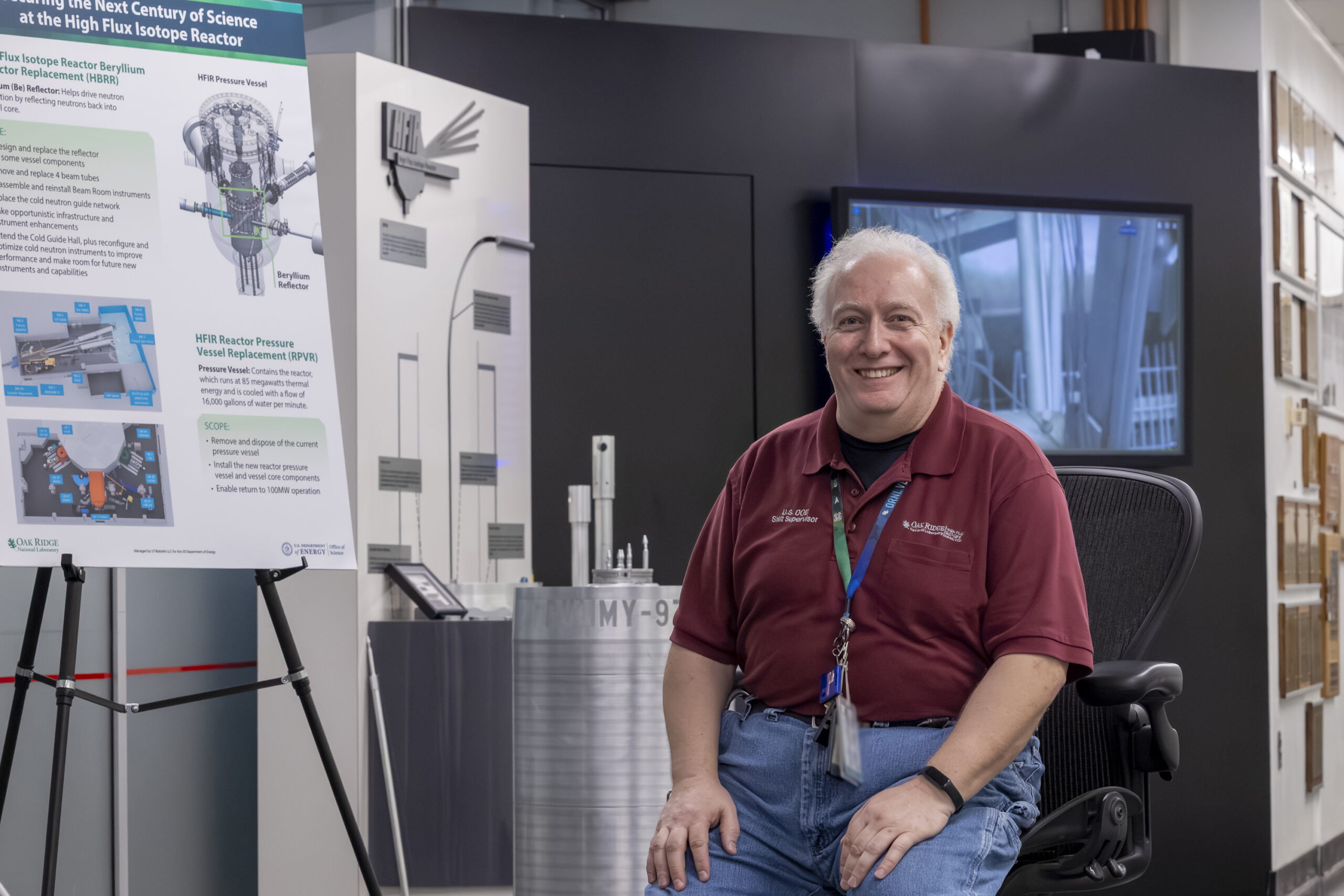 A smiling ORNL employee sits beside a High Flux Isotope Reactor display exhibit and reactor component model.