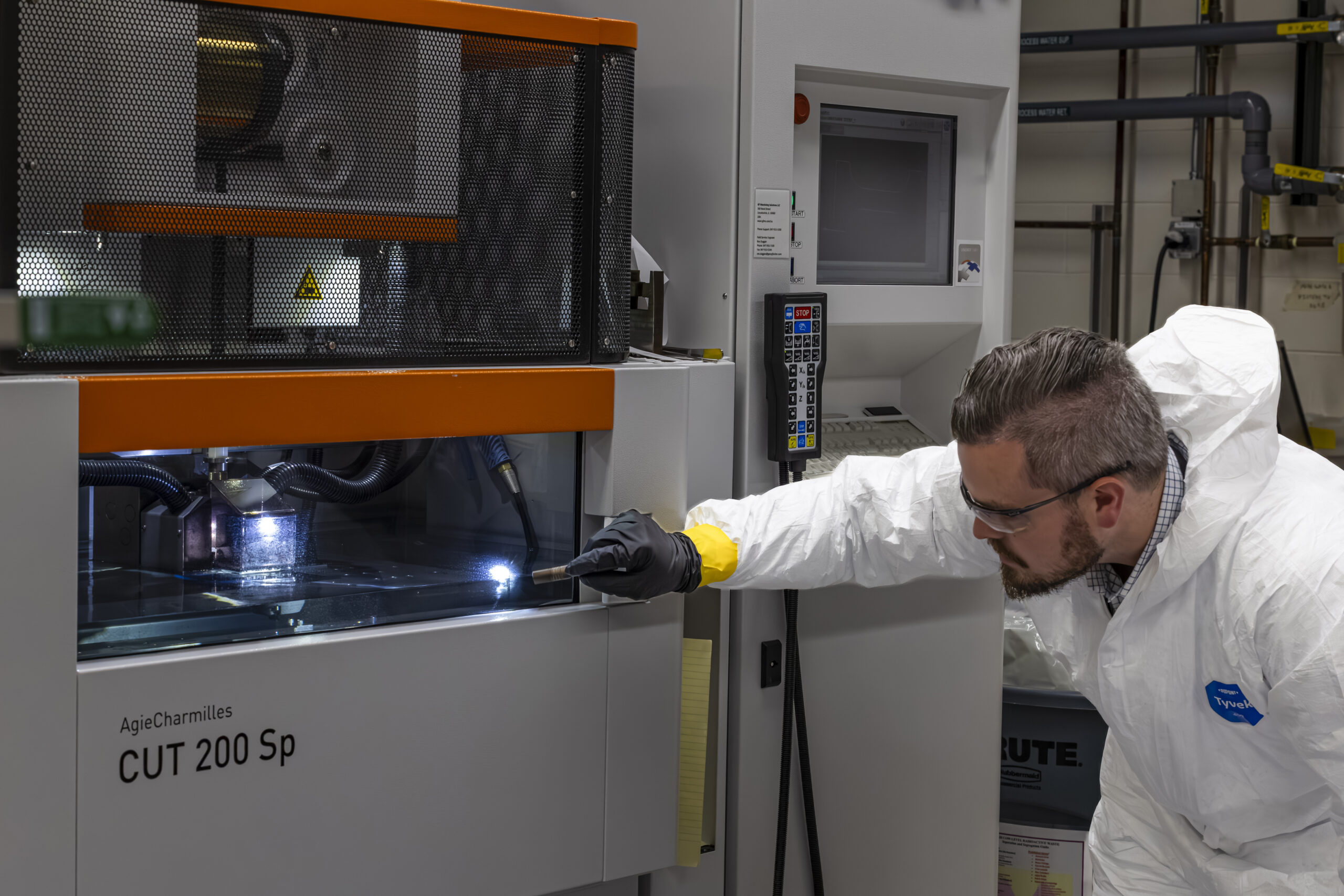 A man in protective lab clothing and safety glasses examines a glowing work area inside an AgieCharmilles CUT 200 Sp machine, using a tool to inspect the component being processed.