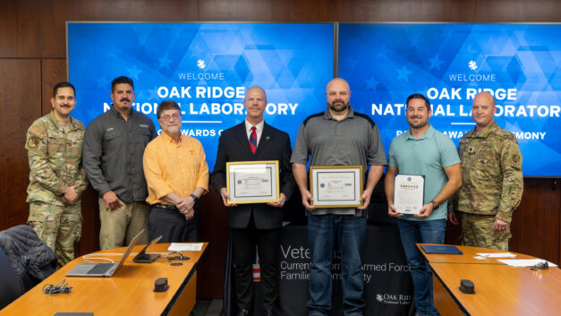 Seven men, including uniformed service members and ORNL staff, stand in front of Oak Ridge National Laboratory Patriot Awards Ceremony screens holding framed awards and a statement of support on February 27, 2026.