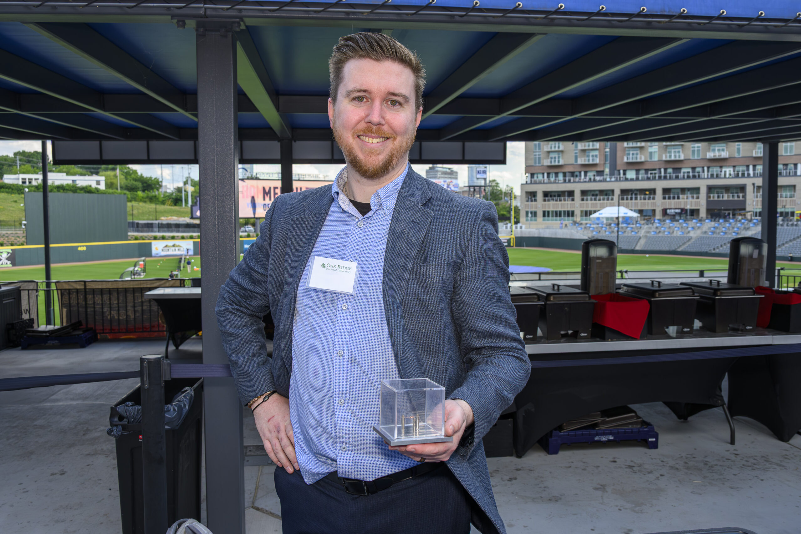 Man in a blazer and collared shirt smiles at the camera while holding a prototype technology, which looks like a cube. He stands on a concourse with a soccer field in the background.