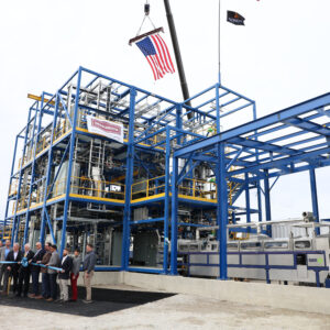 Officials line up to cut a ceremonial ribbon in front of a newly constructed industrial processing unit made of blue structural steel.