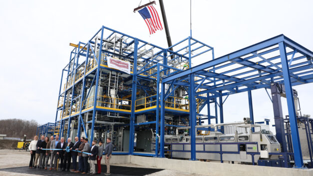 Officials line up to cut a ceremonial ribbon in front of a newly constructed industrial processing unit made of blue structural steel.