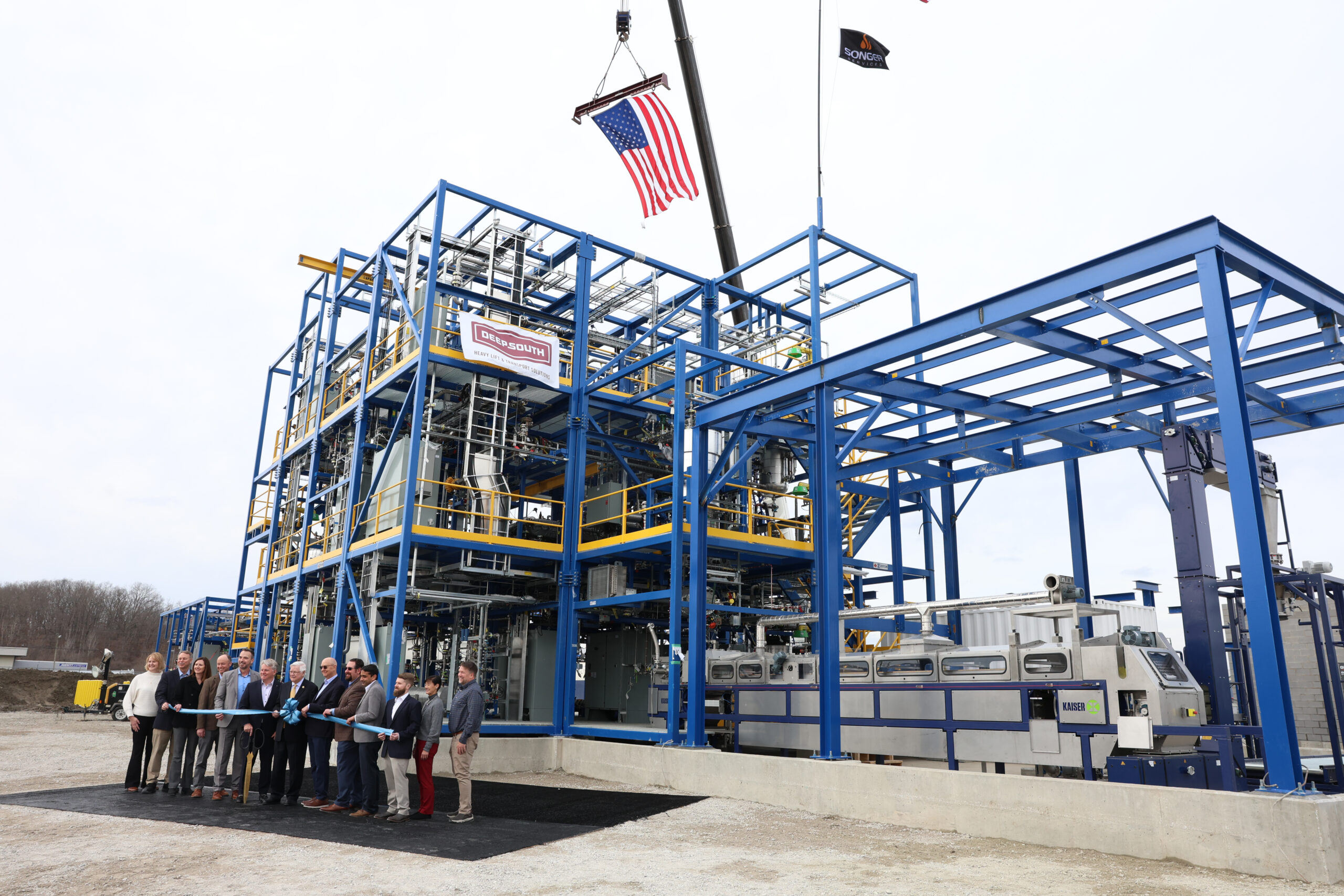 Officials line up to cut a ceremonial ribbon in front of a newly constructed industrial processing unit made of blue structural steel.