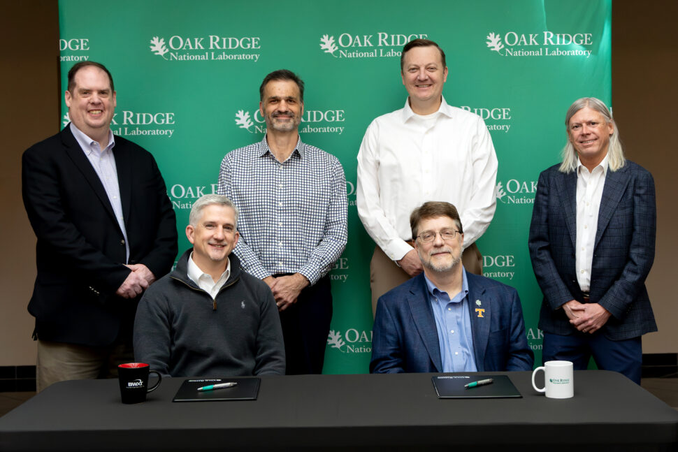 Group photo of six leaders from Oak Ridge National Laboratory and BWXT Nuclear Fuel Services following an MOU signing, with two seated at a table and four standing behind them in front of a green ORNL backdrop.