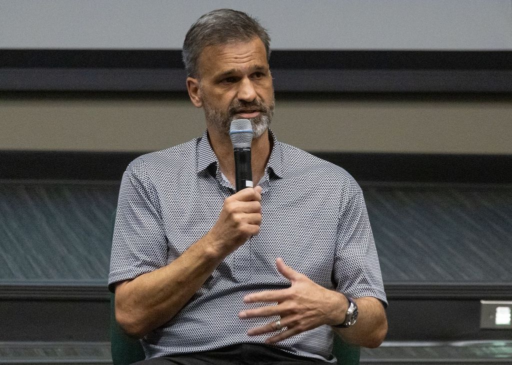 A man with short hair and a trimmed beard speaks into a handheld microphone while seated indoors, gesturing with one hand during a presentation.