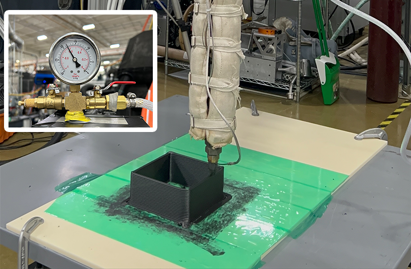 A 3D printing nozzle wrapped in insulation extrudes black composite material into a small square mold on a green and white flat surface in a lab setting. Inset shows a close-up of a pressure gauge connected to brass valves and tubing.