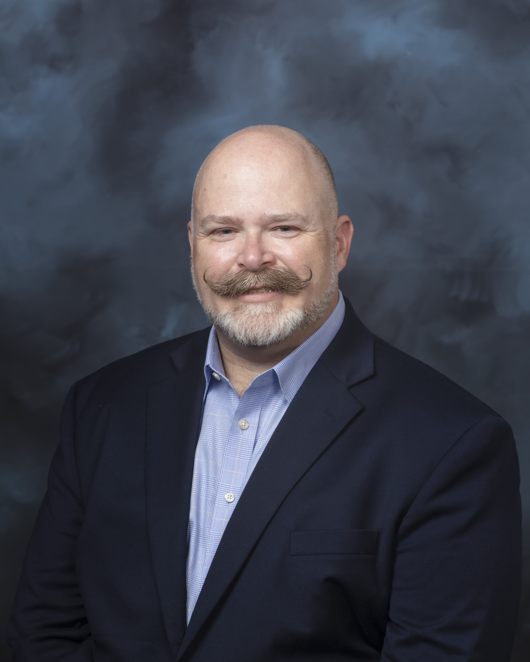 Portrait of a bald man with a handlebar mustache and beard, wearing a dark blue blazer and light blue collared shirt, posing against a blue-gray mottled background.
