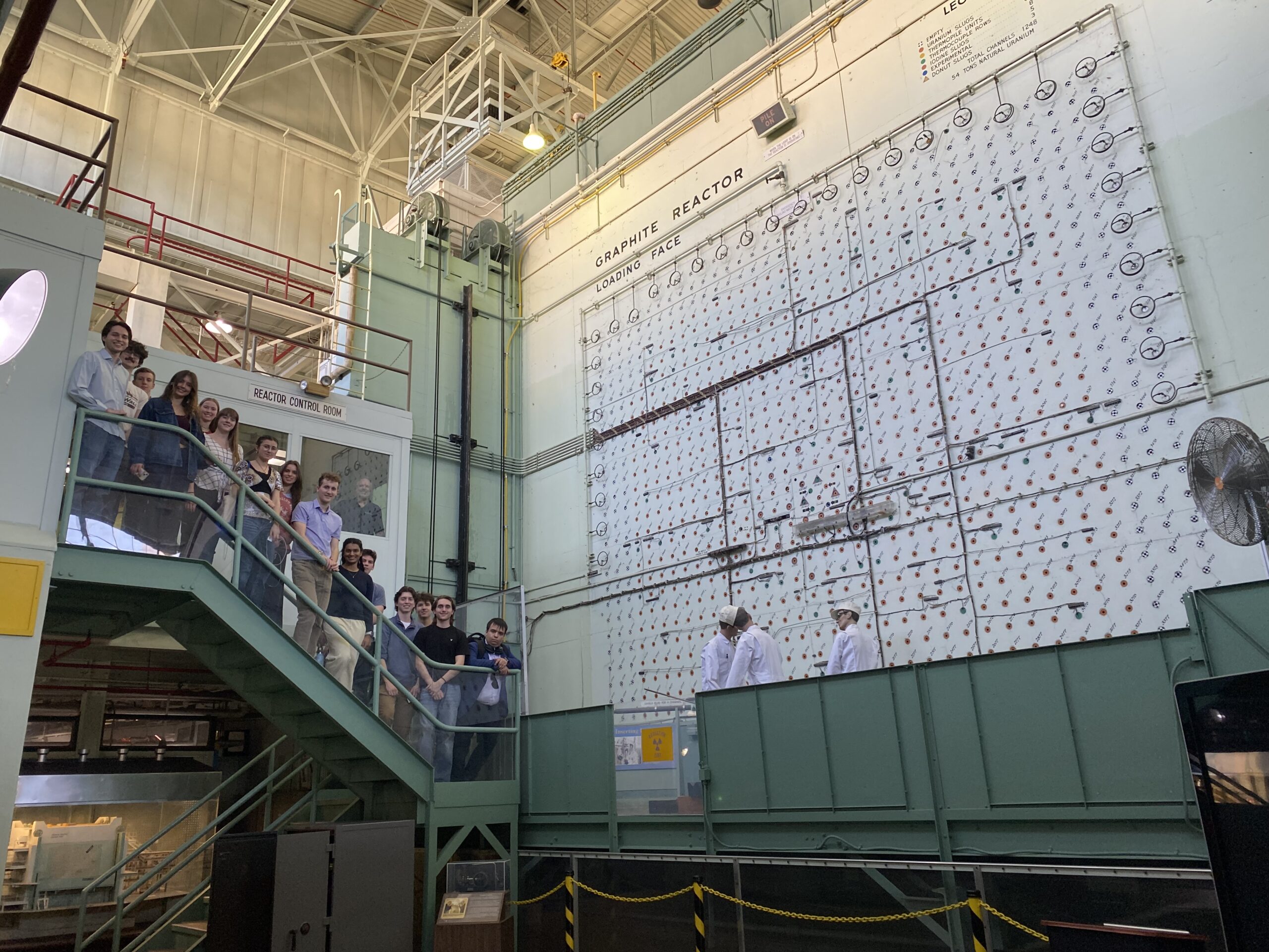 A crowd of college students pose for a photo on the stairs leading to control room in a cavernous industrial building.