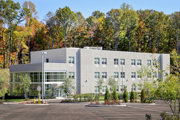 Three-story modern guest house with a light gray exterior, large windows, and a glass-front entrance, surrounded by landscaped shrubs and young trees. An empty parking lot fills the foreground, while a wooded hillside with colorful autumn foliage forms the backdrop under a clear blue sky.