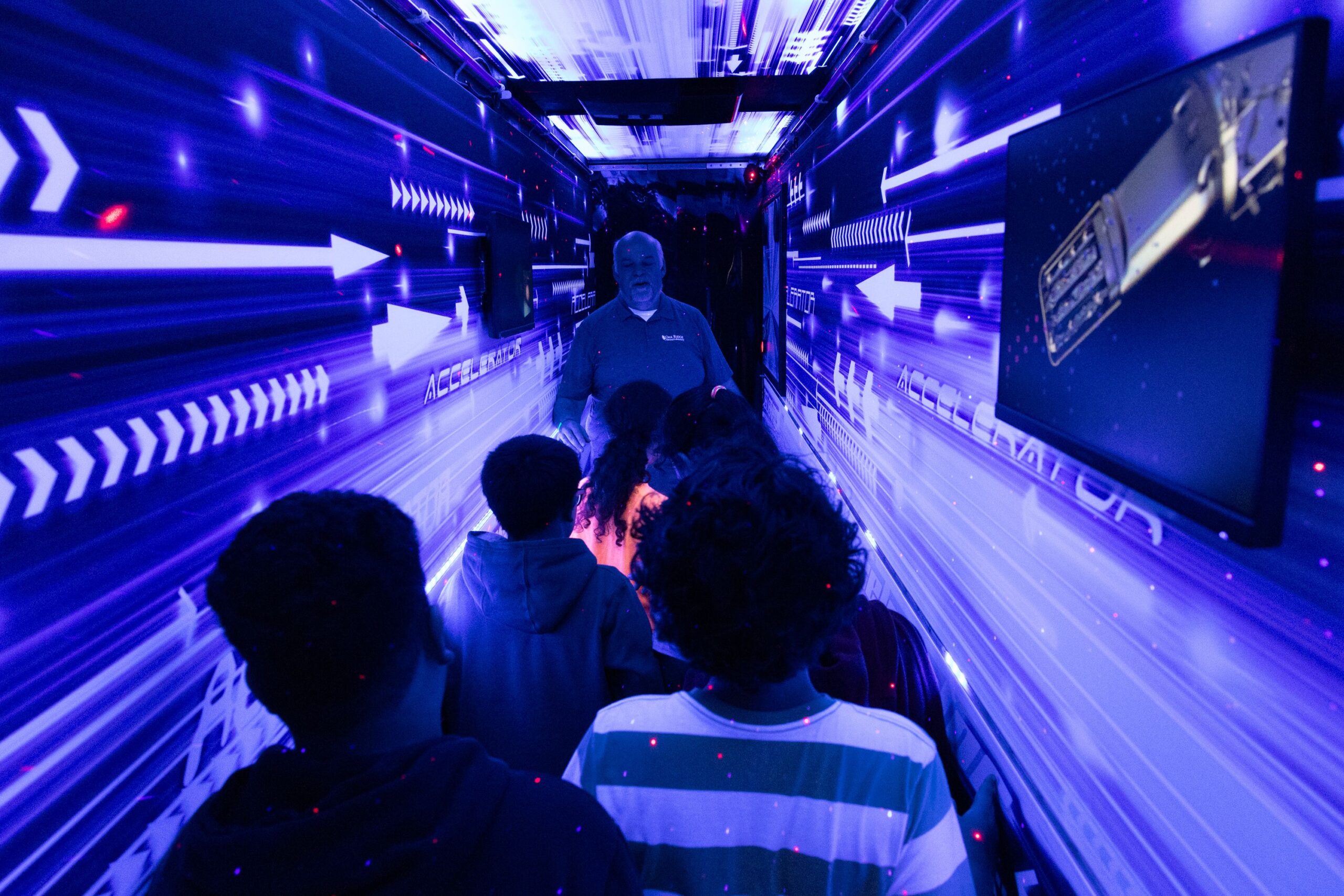 Thomas Proffen leads students through a traveling science fair exhibit, which looks to be lit with black light, giving off a purple hue.