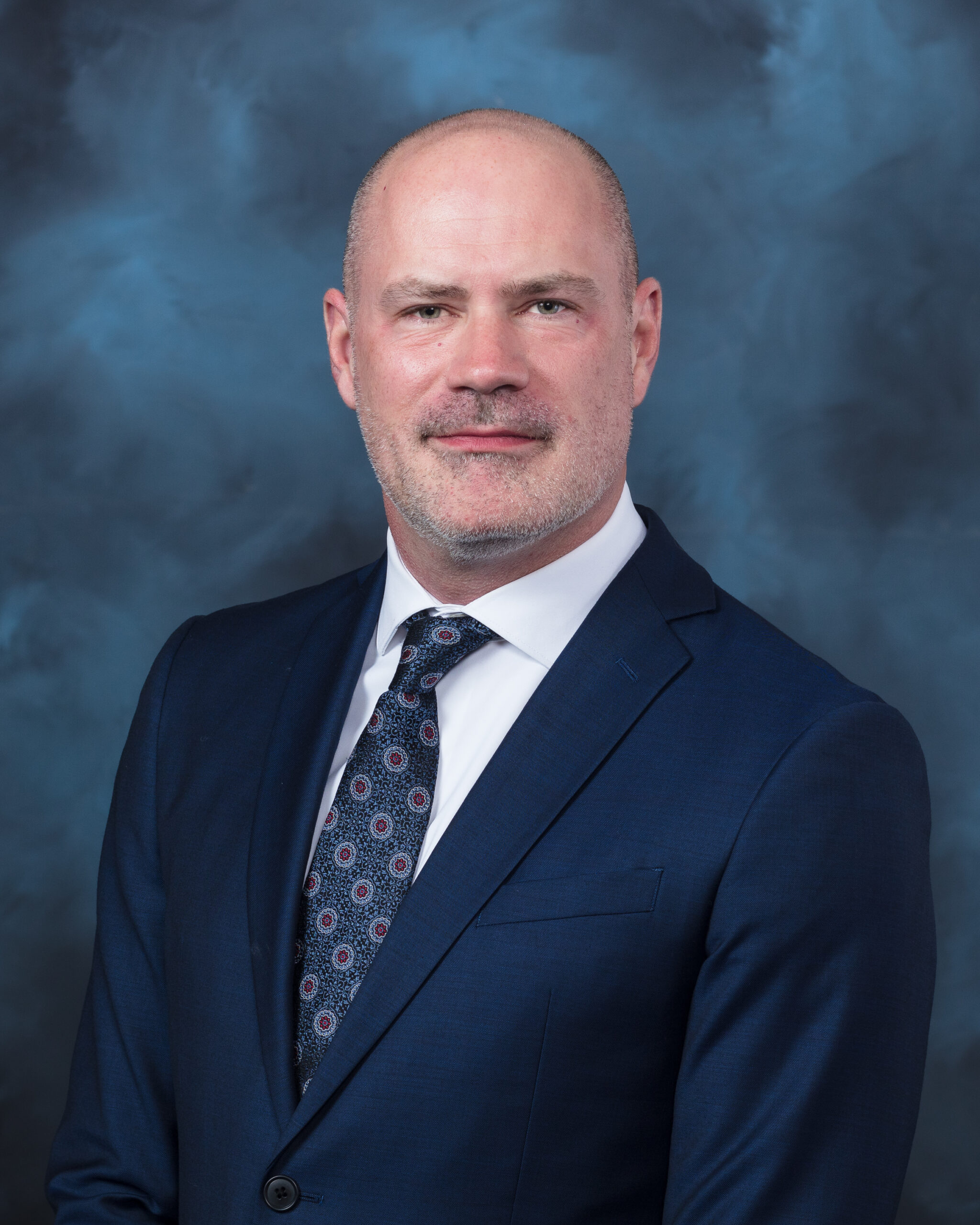 Portrait of a man with short light hair and trimmed beard, wearing a dark blue suit, white shirt, and patterned tie, posed against a blue-gray background