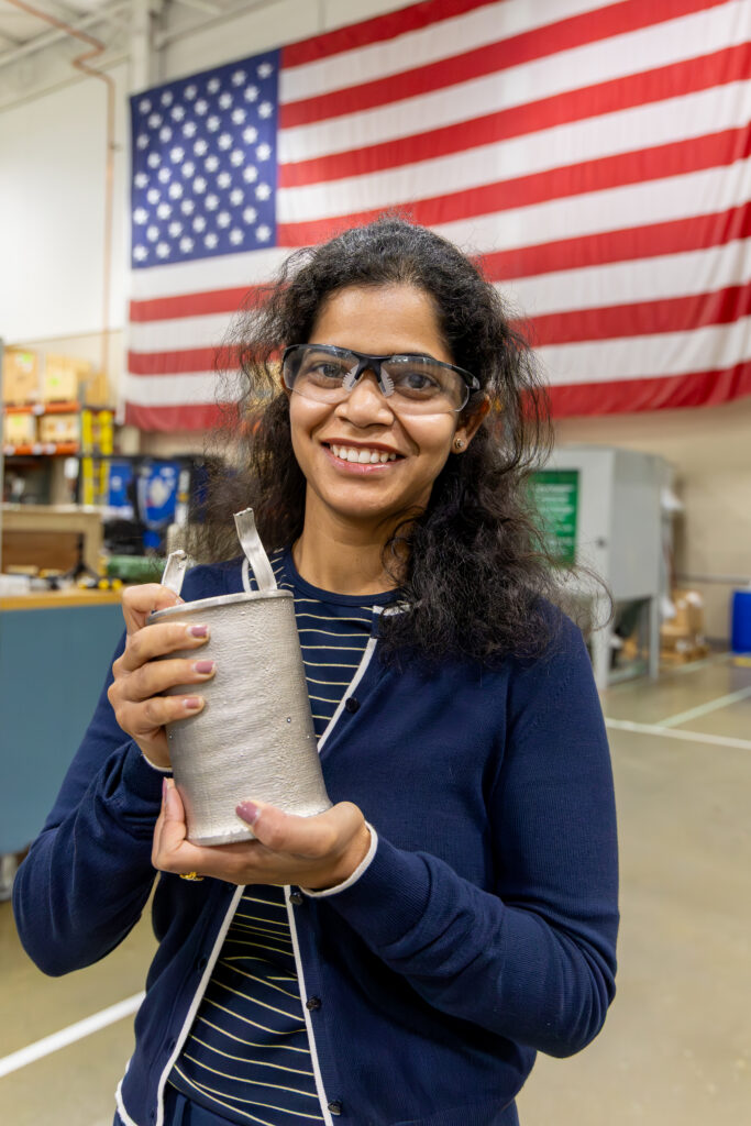 ORNL researcher Vanshika Singh holds an electroformed nickel component developed for advanced nuclear energy manufacturing research while standing in front of a large American flag.