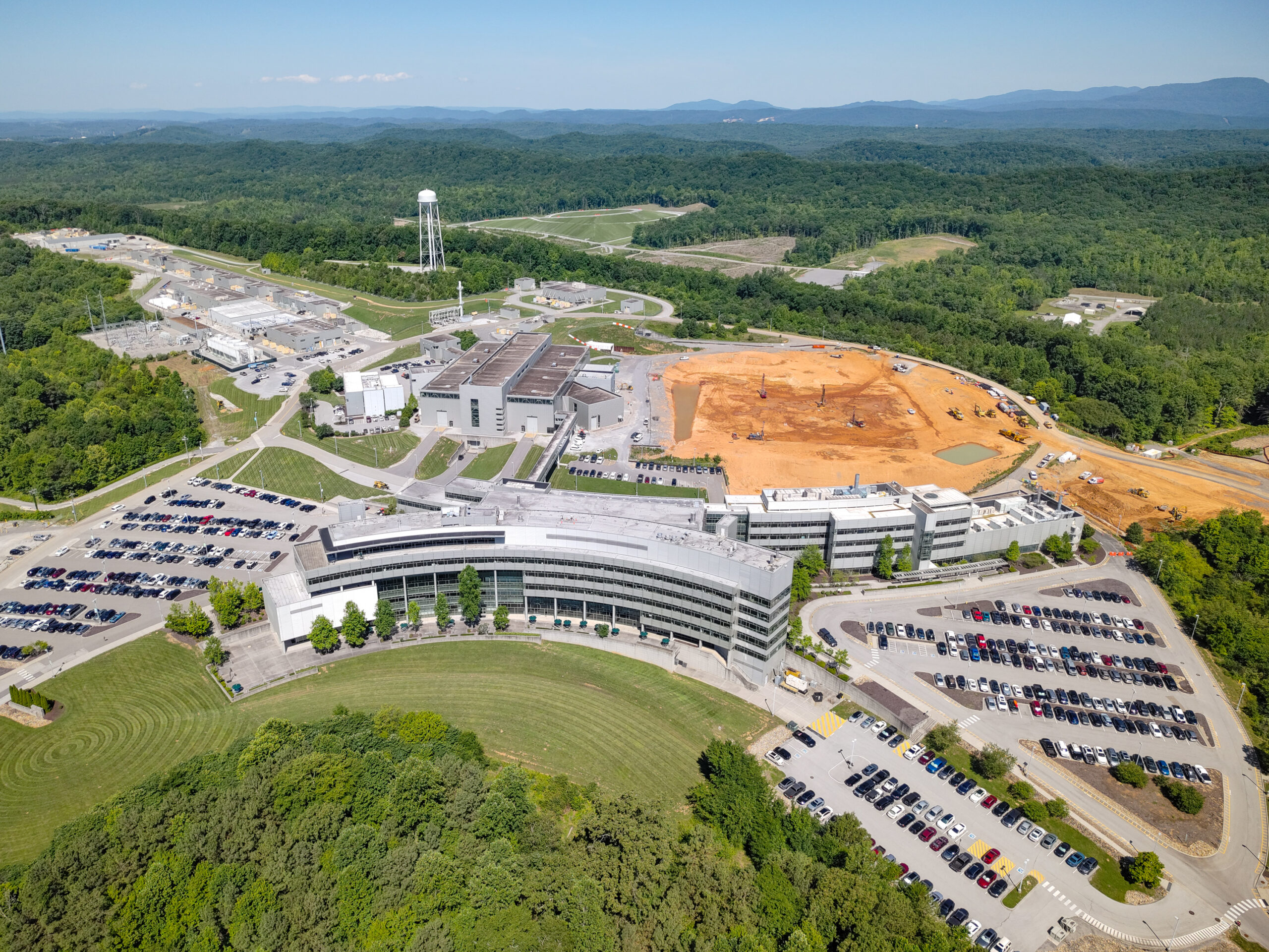 Aerial photographs showing site preparation and early construction for the Second Target Station at the Spallation Neutron Source at Oak Ridge National Laboratory, with cleared land, construction equipment, and nearby research buildings surrounded by wooded hills.
