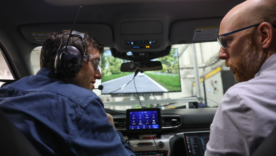 Two researches in safety goggles simulate driving conditions within a lab setting. One researcher is in the driver's seat while looking at the other in the passenger's seat.
