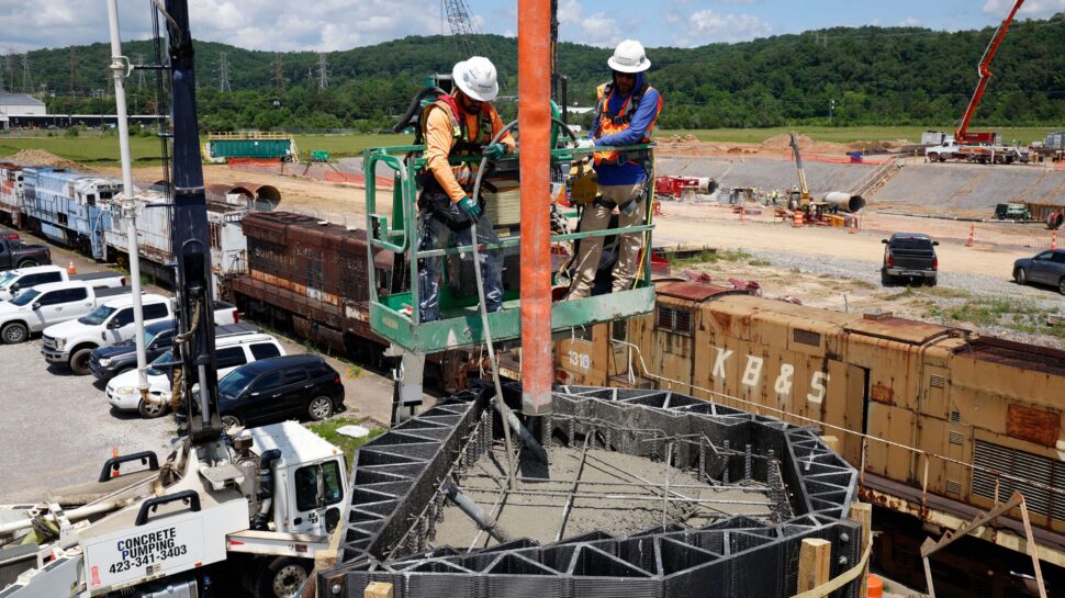 Two men wearing safety equipment like helmets and vests work from a crow's nest on a construction site.