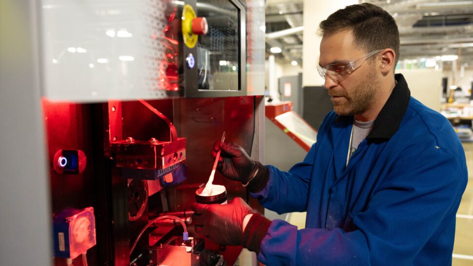 A scientist wearing safety goggles holds illuminated materials in a lab setting.