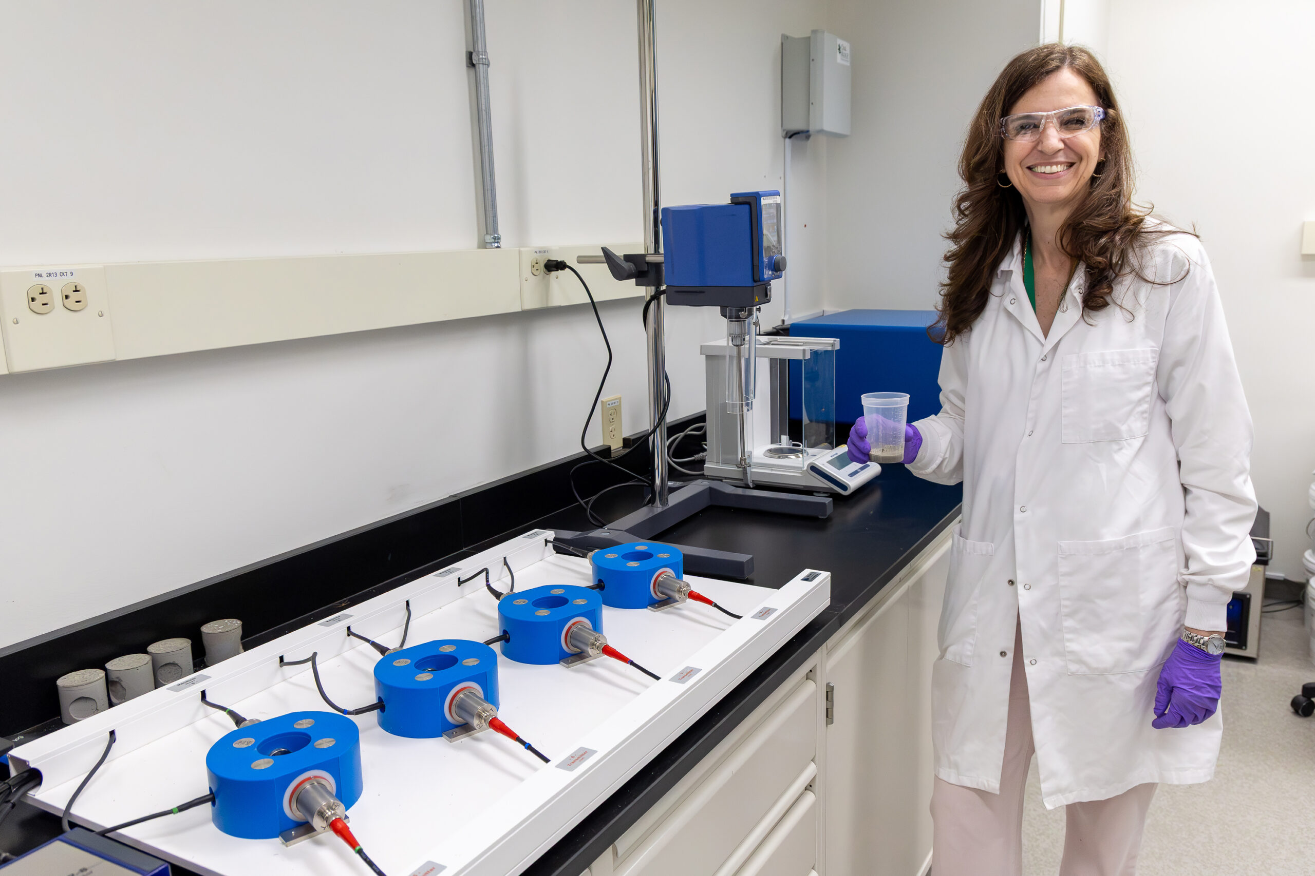Scientist in a lab coat and safety glasses holds a sample container beside laboratory mixing equipment.
