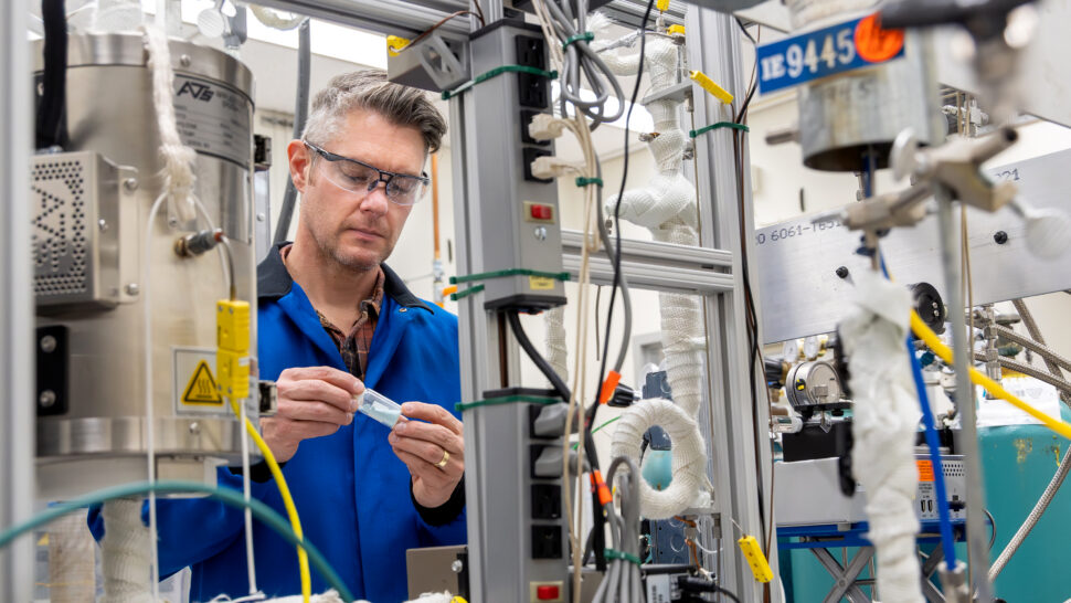 Andrew Sutton, wearing safety glasses and a blue laboratory coat, operates and monitors chemical processing equipment while conducting biomass conversion research in an ORNL laboratory.