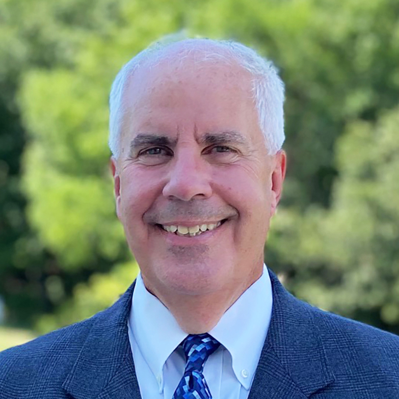 A man in a blue jacket and white shirt looks into the camera in front of a green background