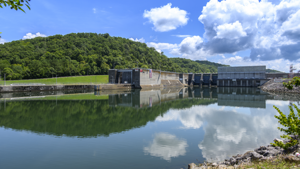 A wide view of Melton Hill Dam reflects in calm water beneath large summer clouds during the Appalachian Hydropower Workshop. The hydroelectric facility is surrounded by forested hills.