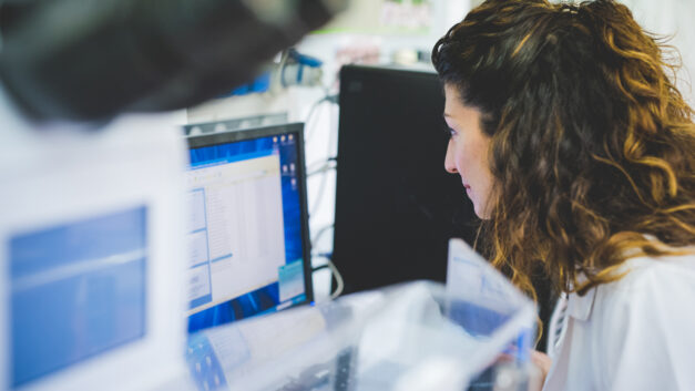 A woman with wavy brown hair, wearing a lab coat, sits in front of a computer in a brightly lit laboratory. She's looking at the screen, which displays data or code. A microscope is visible to her left, slightly out of focus. Image from Envato.