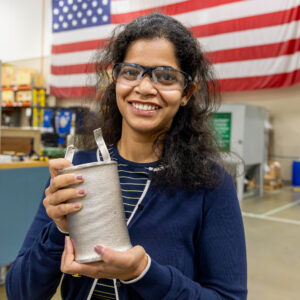 ORNL researcher Vanshika Singh holds an electroformed nickel component developed for advanced nuclear energy manufacturing research while standing in front of a large American flag.