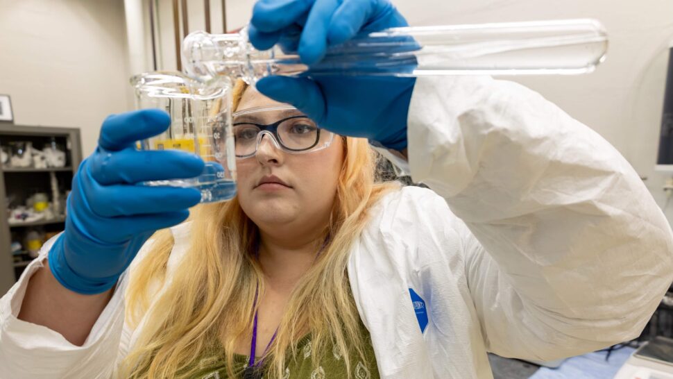 A researcher wearing safety glasses, a protective gown and blue gloves examines a glass tube while holding a flask containing yellow liquid.