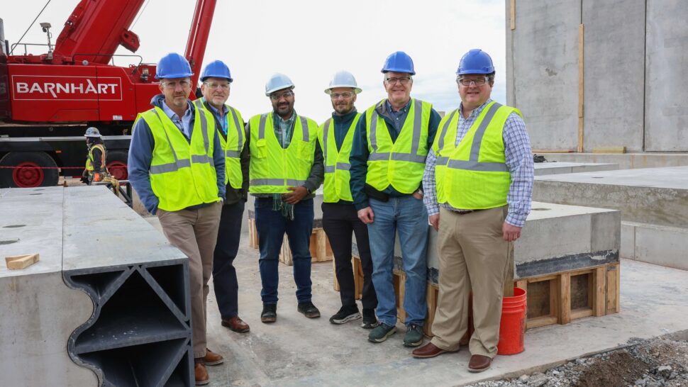 Six people wearing hard hats and high-visibility vests stand among concrete structures at a construction site, with a red crane behind them.