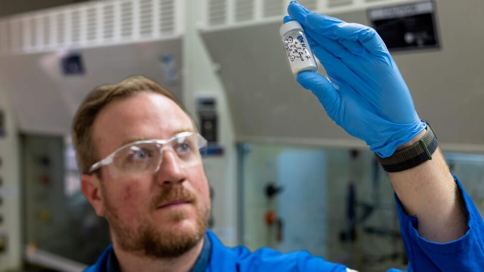 A researcher wearing safety glasses and blue gloves examines a small vial containing a chemical sample in a laboratory.