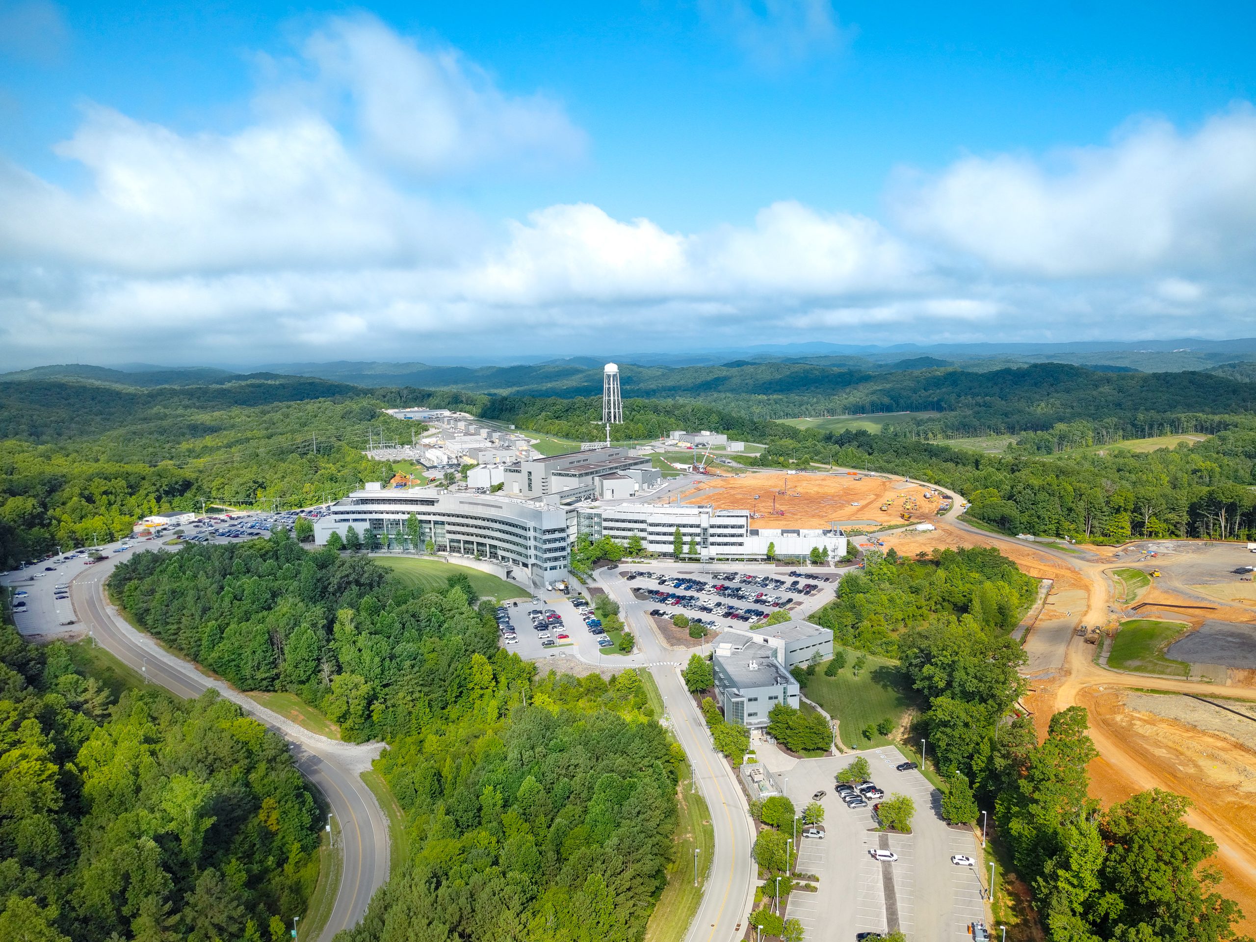 Aerial photograph of the Spallation Neutron Source, Second Target Station construction site, and Center for Nanophase Materials Sciences at Oak Ridge National Laboratory by green forested hills, showing laboratory buildings, parking areas, and nearby construction zones under blue skies with clouds.