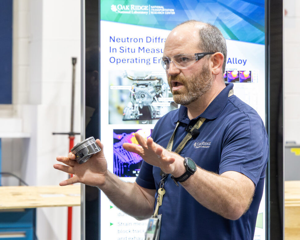 Scott Curran of ORNL speaks to a group of Industry Summit attendees inside a transportation research laboratory while holding engine components during a technical presentation.