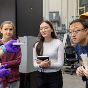 Neutron scattering scientist Lisa Debeer-Schmitt holds up a small sample mount as students gather closely around her during hands-on instruction at the GP-SANS instrument.