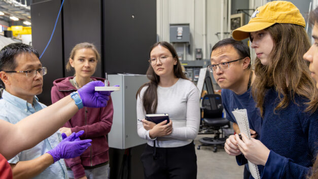 Neutron scattering scientist Lisa Debeer-Schmitt holds up a small sample mount as students gather closely around her during hands-on instruction at the GP-SANS instrument.