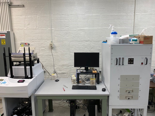 A rectangular cabinet-like device on legs sits to the right of a desk with a small, elevated monitor; to the left of the desk, a small printer-like machine sits behind four tiny black columns