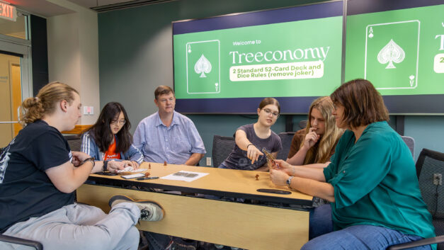 Erin Webb, Robin Clark, and students sit around a table learning and playing the educational strategy card game Treeconomy during the Appalachian Regional Commission Science Institute, with game instructions displayed on monitors in the background.
