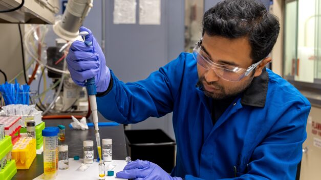 A man in a blue lab coat injects chemicals into a test tube.